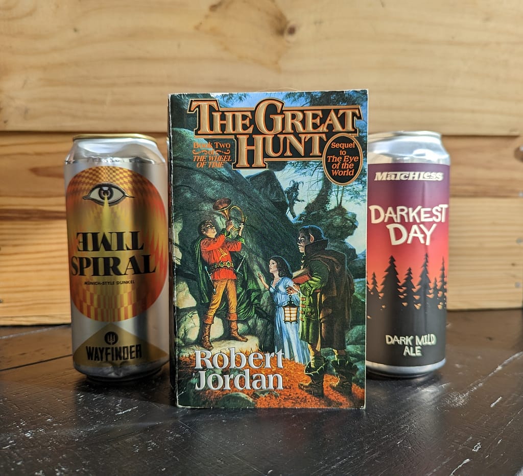 Paperback copy of The Great Hunt by Robert Jordan in the foreground, with a can of Time Spiral beer by Wayfinder and a can of Darkest Days beer by Matchless Brewing. All items are on a black table with a wooden crate in the background.