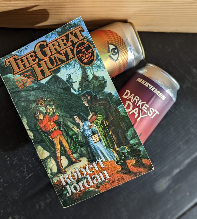 Paperback copy of The Great Hunt by Robert Jordan lays on top of two cans of beer. All items lay flat on a black table with a wooden crate in the background.