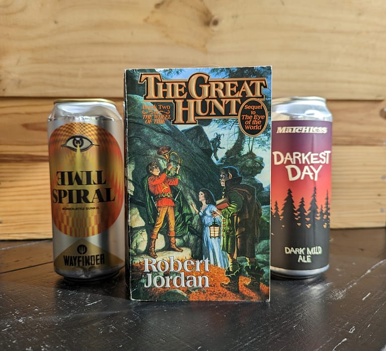 Paperback copy of The Great Hunt by Robert Jordan in the foreground, with a can of Time Spiral beer by Wayfinder and a can of Darkest Days beer by Matchless Brewing. All items are on a black table with a wooden crate in the background.